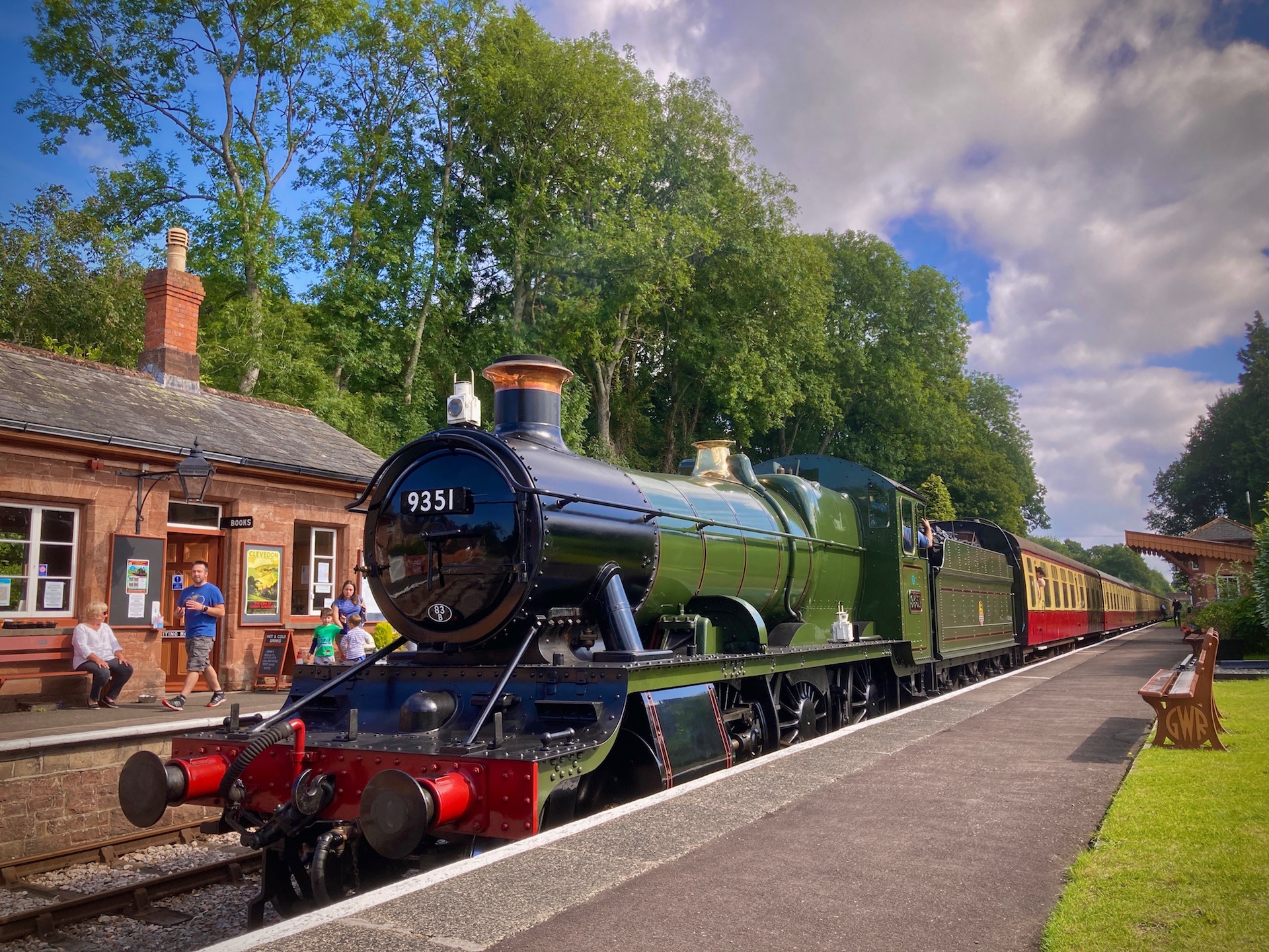 A steam engine waits alongside an empty platform at Crowcombe Station on the West Somerset Railway. On the far side of the track the restored Victorian station buildings can be seen.