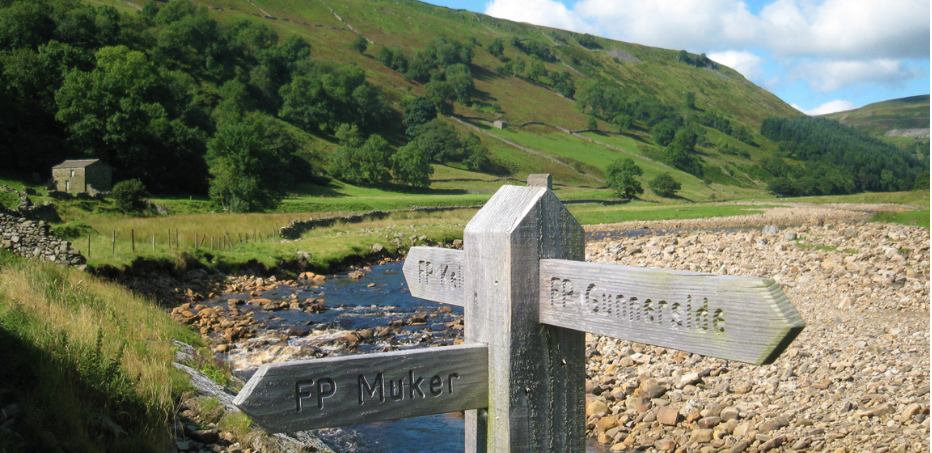 A Yorkshire Dales wooden signpost stands in front of a meandering river with a shingle beach. Beyond the river is a steep, partially tree-clad fell.