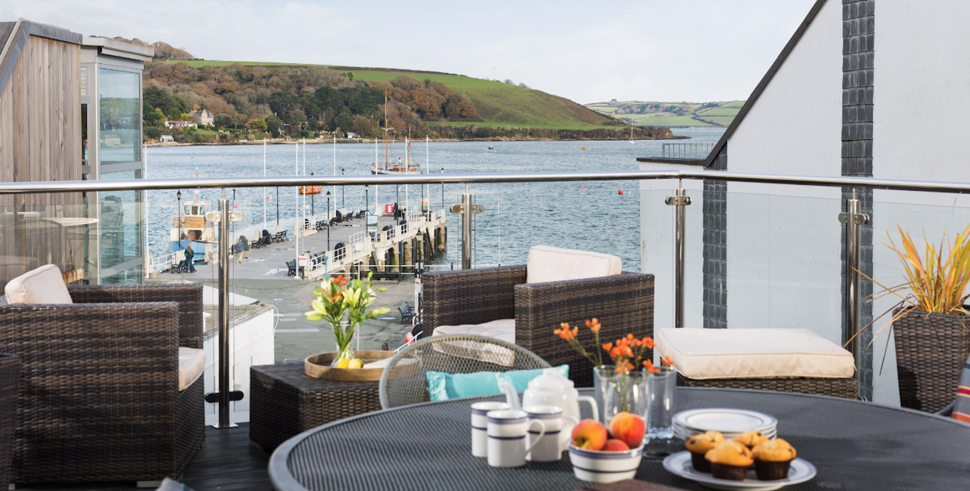 a table with a fuit bowl and wine glasses on a deck with wooden chairs overlooks Falmouth harbour, its small pier and the sea.