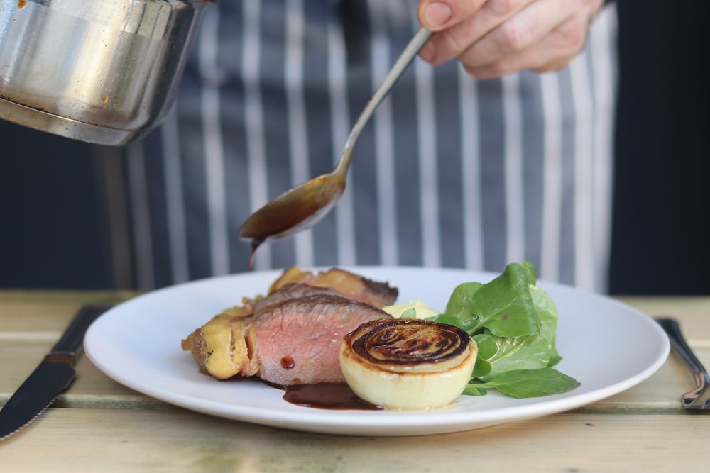 A chef in striped apron pours a jus on a rare steak on a plate.