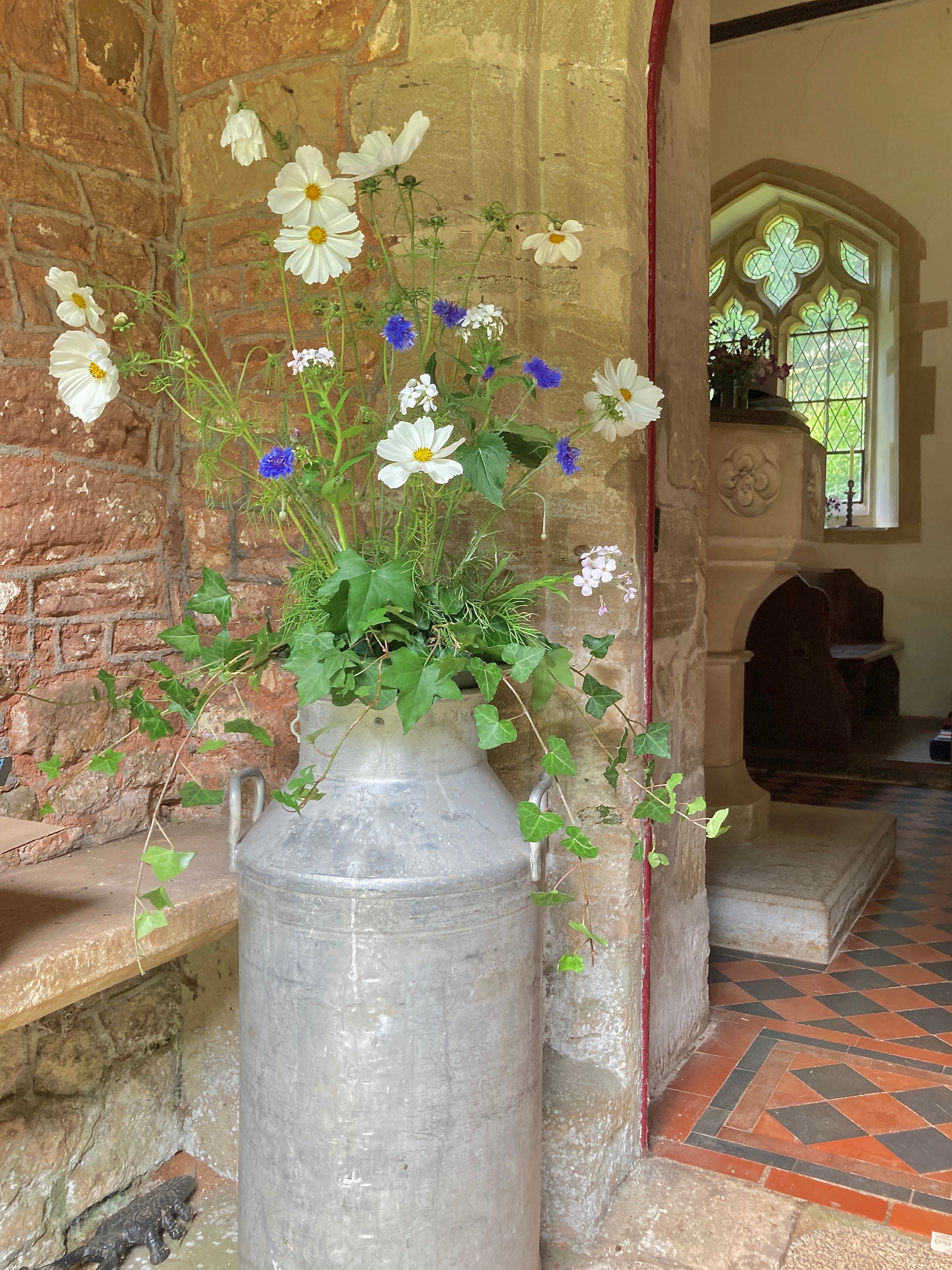 An old milk churn filled with flowers stands in a Somerset church porch.