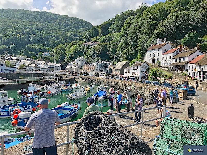 Lynmouth harbour full of pleasure and fishing boats. In the foreground is a stack of lobster pots. Little coottages and shops line the harbour edge behind a steep backdrop of trees.