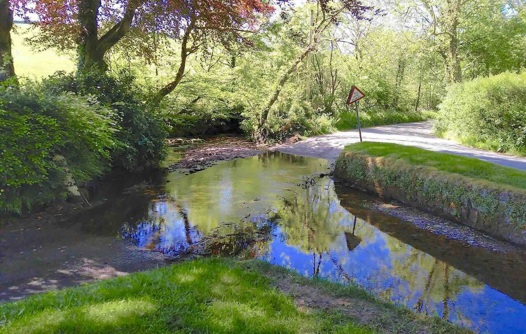 A tree-lined country lane runs through an Exmoor river ford.