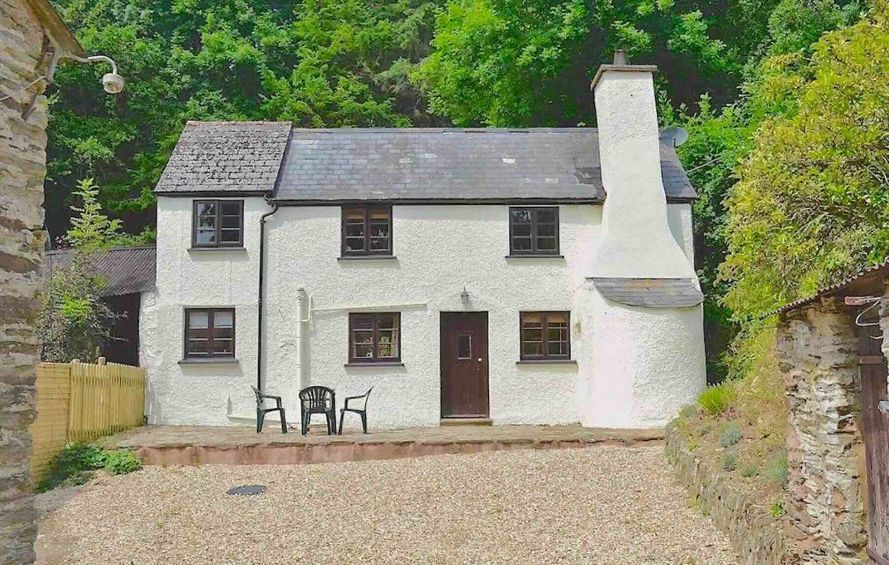 Exterior of a slate-roofed Exmoor holiday cottage with a large chimney and bread oven stack, overlooked by tall woodland trees.