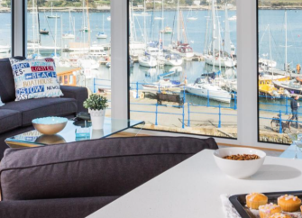 An open plan lounge with worktop and large comfortable sofas overlooks a harbourside view in Falmouth through floor-to-ceiling windows.