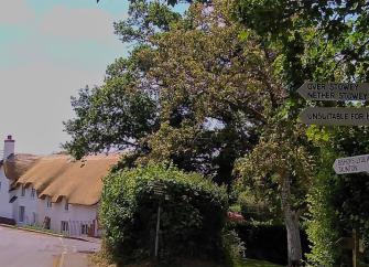 Crowcomba village street. In the foreground is a signpost with directions to local villages. Behind it are trees and a newly thatched row of cottages.