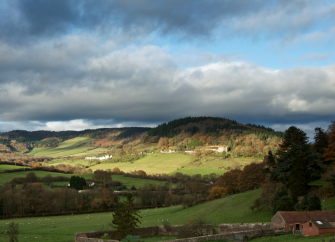 A landscape view of the Wye Valley with trees and open fields. In the foreground are some stone farm buildings  beyond which the land rises to a small hill on the horizon.