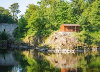 a wooden lodge in Aberdeenshire backed by trees is reflected in the calm waters of a river below it.