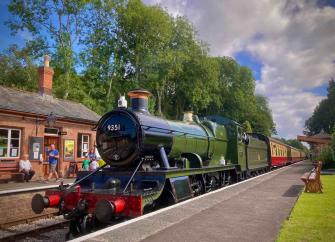 A restored steam engine and carriages stands alongside a platform  in a small village railway station in a rural setting.