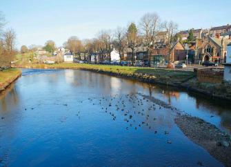 Town houses and shops overlook a wide, shallow river bordered by trees in winter.