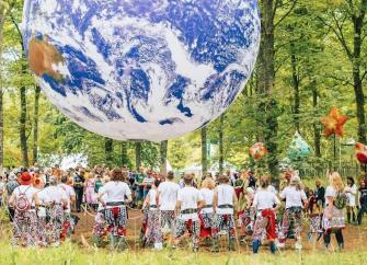 Festival goers in garish attire admire a large inflatable globe hanging from woodland trees in Kendal.