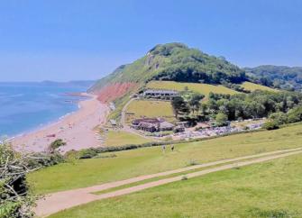 Branscombe beach in Devon. A field slopes down to a cluster of sea shanties overlooking a wide shingle beach, beyond which the lad rises to form cliffs topped by woodland. 