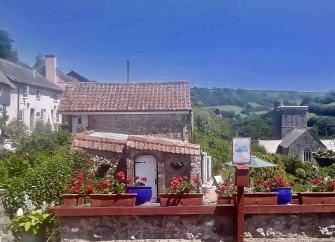 A view down the valley in Branscombe in East Devon. In the foreground is a large trough of flowers, behind which are cottages and a church tower. To either side are steep fields and woodland.