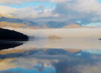 In the Lake District, a calm surface of a large lake reflects the fleecy cloud-filled sky above