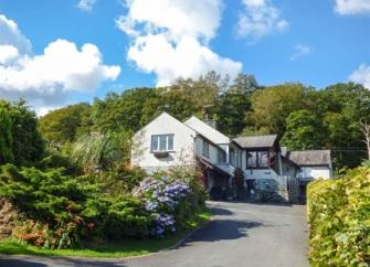 A large Coniston holiday home at the head of a hedge-lined tarmac drive with a mature woodland background.