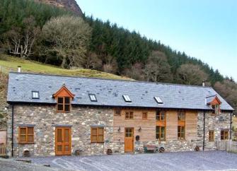 A slate-roofed, stone holiday cottage in Powys nestles beneath a step hill toppped with a wood.