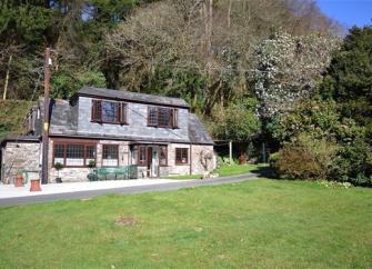 A 2-storey double-fronted stone-built holiday cottage in Cornwall with a slate roof overlooks a large lawn. Behind the cottages is a bank of mature trees in full leaf.