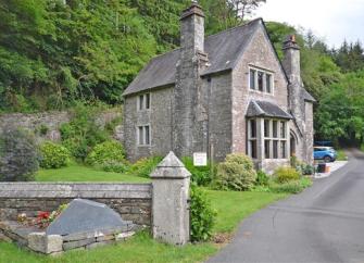 A drive leds to a tall stone house with a mullioned bay window. A wood grows behind the house.