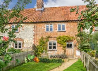 Exterior of a quaint stone-built Norfolk holiday cottage overlookings. mature garden and lawn.