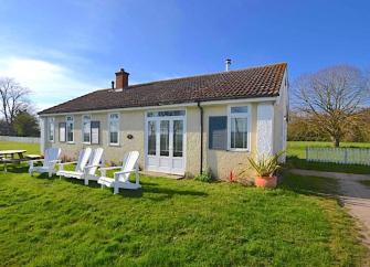 Exterior of an Osea Island holiday bungalow surrounded by lawns containing sun loungers and a picnic table.