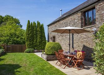 A table  with parasol and chairs tands on a sunny patio behind a stone built holiday cottage in Devon.