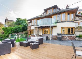A wooden deck with lounge furniture sits in front of a contemporary 3-storey house.