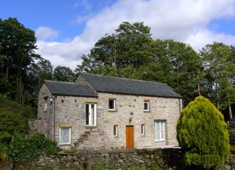 Doiuble-fronted exterior of a stone holiday cottage in Cumbria surrounded by mature trees.
