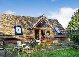 A wood and slate Cotswold holiday cottage overlooks a terrace with dining table and lawn.