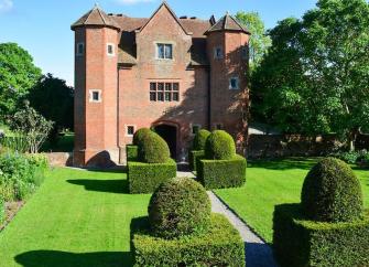 A grand 3-storey, brick-built Tudor house overlooks a large, formal lawned gardens.
