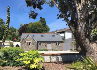 Between shrubs and a mature tree, stands a Cornish 2-storey stone-barn conversion