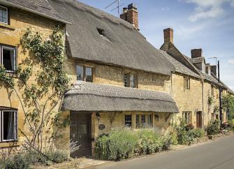 A thatched Cotswold Cottage with a bay window overlooks a village street.