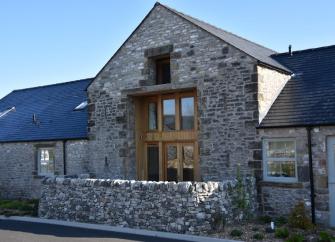 Exterior of a stone-built barn conversion with floor-to-ceiling windows behind a low stone wall.