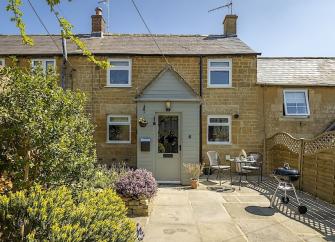 Stone exterior of a terraced Cotswold cottage with a paved garden having a table and chairs.