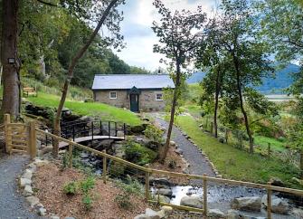 A single storey, stone cottage overlooks a garden with a small stream trickling under a bridge in the foreground.