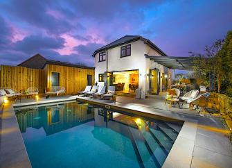 A large, brightly lit house overlooks a swimming pool at dusk.
