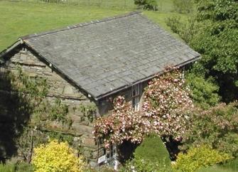 A stone-built, slate-roofed Lake District holiday cottage surrounded by shrubs and small trees with hilly fields in the distance.