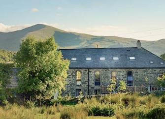 A stone barn conversion backed by mountains overlooks a field with small trees.