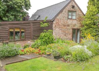 The exterior of a stone-built cottage with a wooden extension overlooks a flower-filled garden.