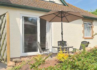 The exterior of a chalet bungalow with a courtyard containing a table with a parasol and chairs.