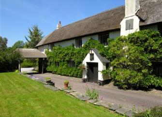 A large, thatched, wisteria-clad Somerset holiday cottage with a covered porch.