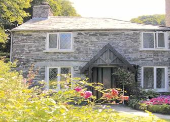 A double-fronted stone cottage overlooks a sunny lawn edged with flowers.