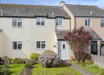 A gable-windowed holiday cottage faces a large lawn bordered by bushy shrubs.