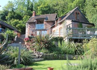 Against a woodland backdrop, a large cottage with a spacious deck overlooks a flower and shrub-filled garden surrounding a lawn.