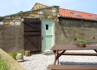 Entrance to a single storey seaside barn conversion in front of which is a wooden picnic table.