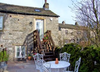 A stone barn conversion with steps to a 1st floor enntrnce from a patio with table and chairs.