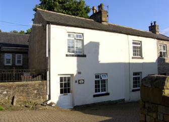 A white-rendered 2-tory, semidetached holiday cottage in Cumbbria overlooks a quiet lane.