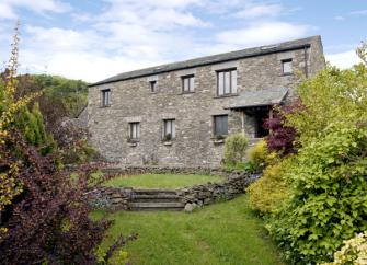A large barn conversion in KEndal overlooks a  lawn surrounded by mature shrubs and bushes.