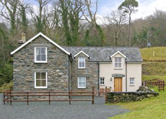 A large slate roofed Lake district holiday cottage its in front of a line of tall tres.