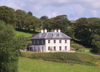A grand 3-storey Georgian country house in the Cornish countryside partly surrounded by trees.