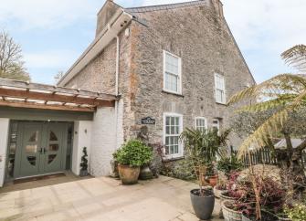 A tall, 2-storey, stone-built cottage overlooking a mature garden with a terrace and palms.